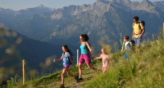 Familie mit Kindern wandert auf einem Bergpfad mit Alpenpanorama im Hintergrund.