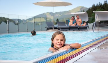 Lachendes Mädchen im Pool mit Bergblick und Liegen im Hintergrund.
