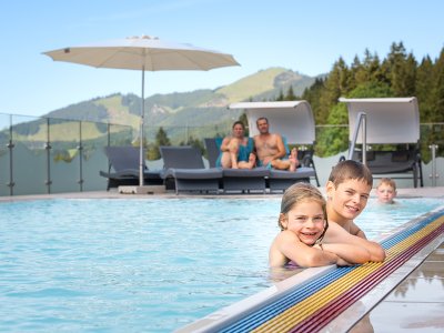 Two smiling kids at the pool edge with mountains and loungers behind.
