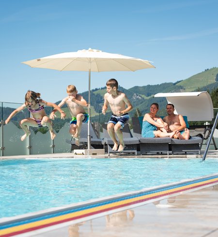 Drei Kinder springen ins Becken, Eltern entspannen auf Liegen.