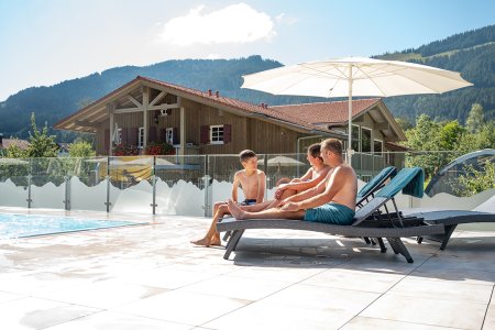 Familie entspannt auf Liegen am Pool vor alpenländischem Haus.