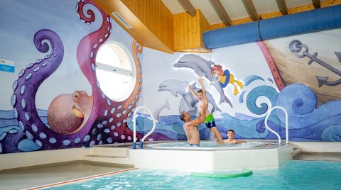 Familie spielt im Hallenbecken mit bunter Unterwasser-Wandmalerei.