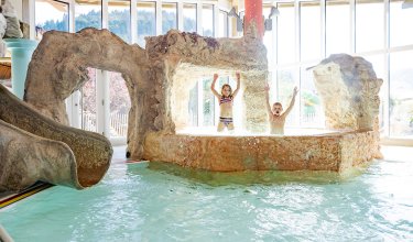 Kinder spielen unter Wasserstrahlen im Hallenbad mit Felsengrotte.