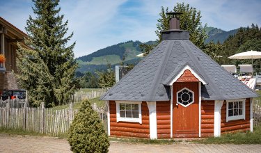 Kleine rote Blockhütte mit schwarzem Dach und Bergblick im Alpenstil.
