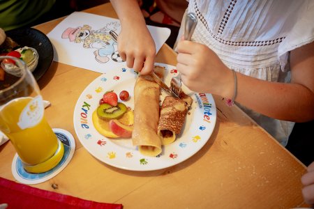 Kind schneidet Palatschinke auf buntem Teller mit frischem Obst und Malbild.