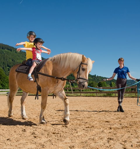 Zwei Kinder reiten zusammen auf einem Pony und machen eine Übung, während die Betreuerin das Pony führt.