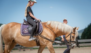 Mädchen reitet ein hellbraunes Pferd, geführt von lächelnder Frau.