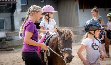 Mädchen sitzt auf Pony, wird von Frau geführt und Kind gehalten.