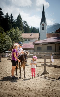 Child on pony led by woman, another child watching nearby.
