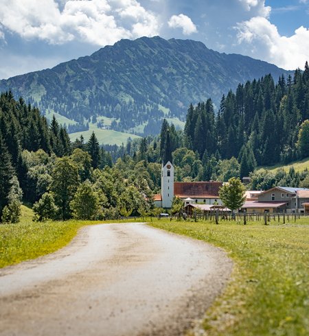 Weg durch grüne Wiese Richtung Dorf mit Kirche und Bergen.